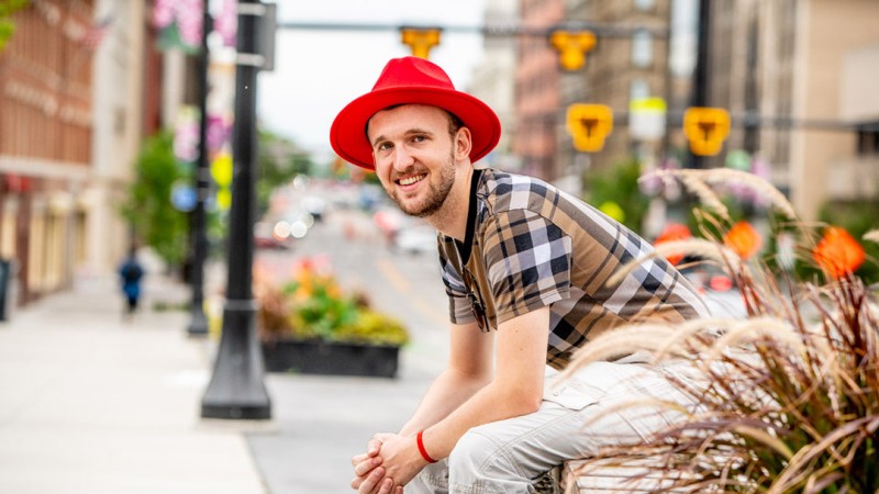 main in short sleeve plaid shirt and red fedora sits smiling at camera