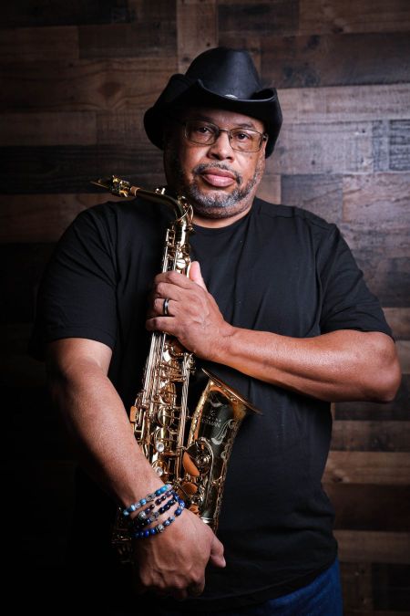 Jimmy Highsmith Jr stands in front of a wood wall holding a saxophone wearing a black cowboy hat and shirt