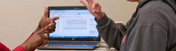 Photo of hands signing, with cochlear implant settings screen on laptop on table behind them