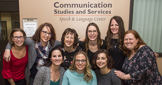 Photo of speech and language center group in front of sign that says Communication Studies and Services Speech and language Center; shows 8 women in a circular format with their arms around each other