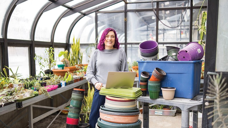 Sustainability Ph.D. student Katie Malarkey ’04 (physics) is weraring a multicoliored sweater and stands in a greenhouse surounded by plants.
