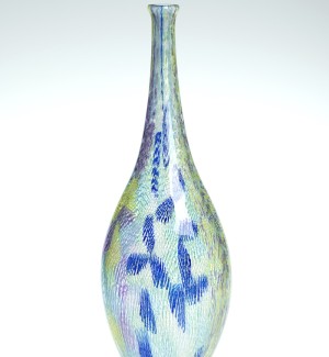 a tall narrow glass bottle form with a clear glass base and multi colored hatching of green and blue forming a pin wheel pattern in places.