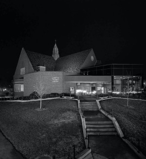 a black and white photograph of a nightime view of a brick building with peaked roof line and 'Lorette Wilmot Library on the wall.