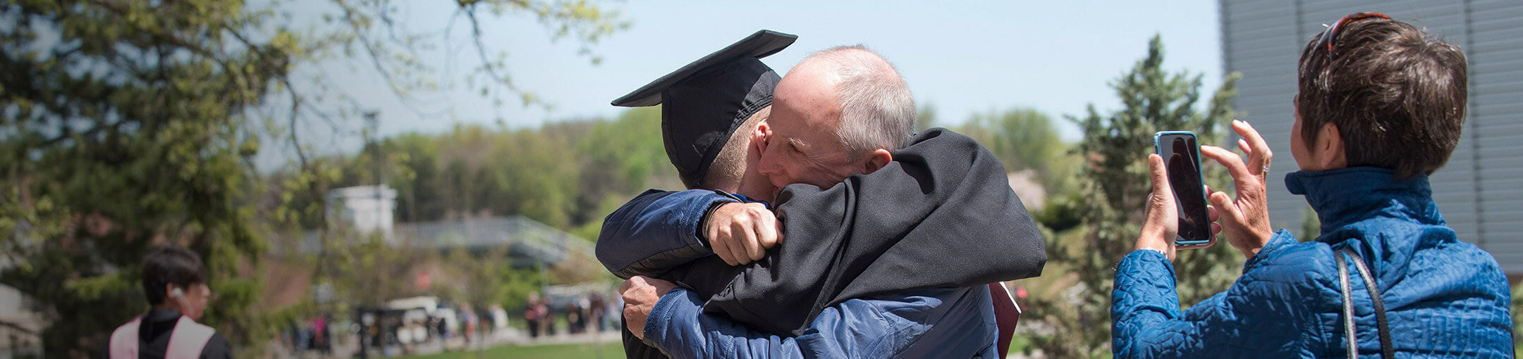 A father hugging his son after graduation while the mother takes a picture
