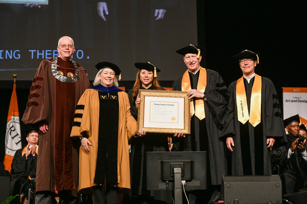 five people posing for photo wearing graduation regalia and holding a framed diploma.
