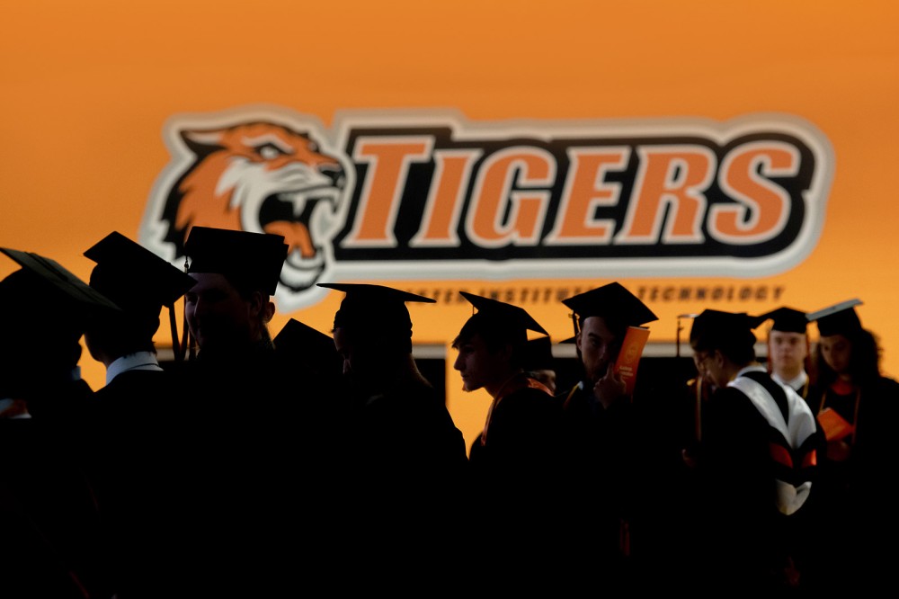 outline of graduating college students wearing mortarboards against an orange R I T Tigers athletics logo in the background.