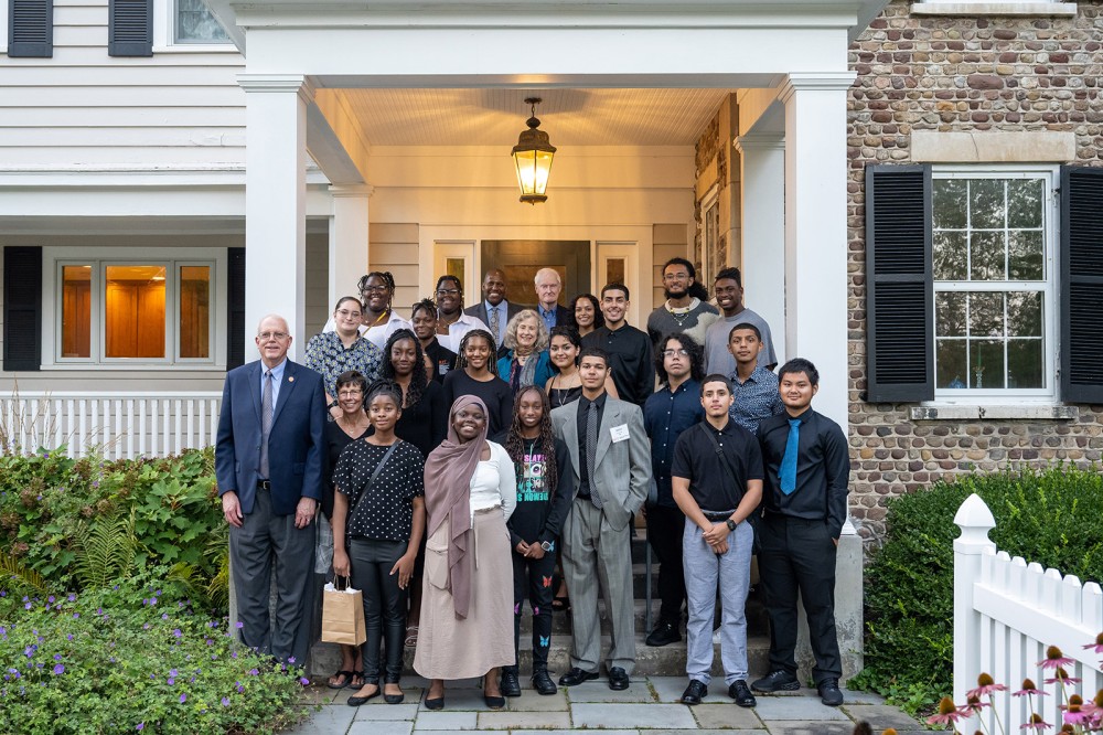 a group of multicultural college students standing in front of a house with two college presidents and their spouses.