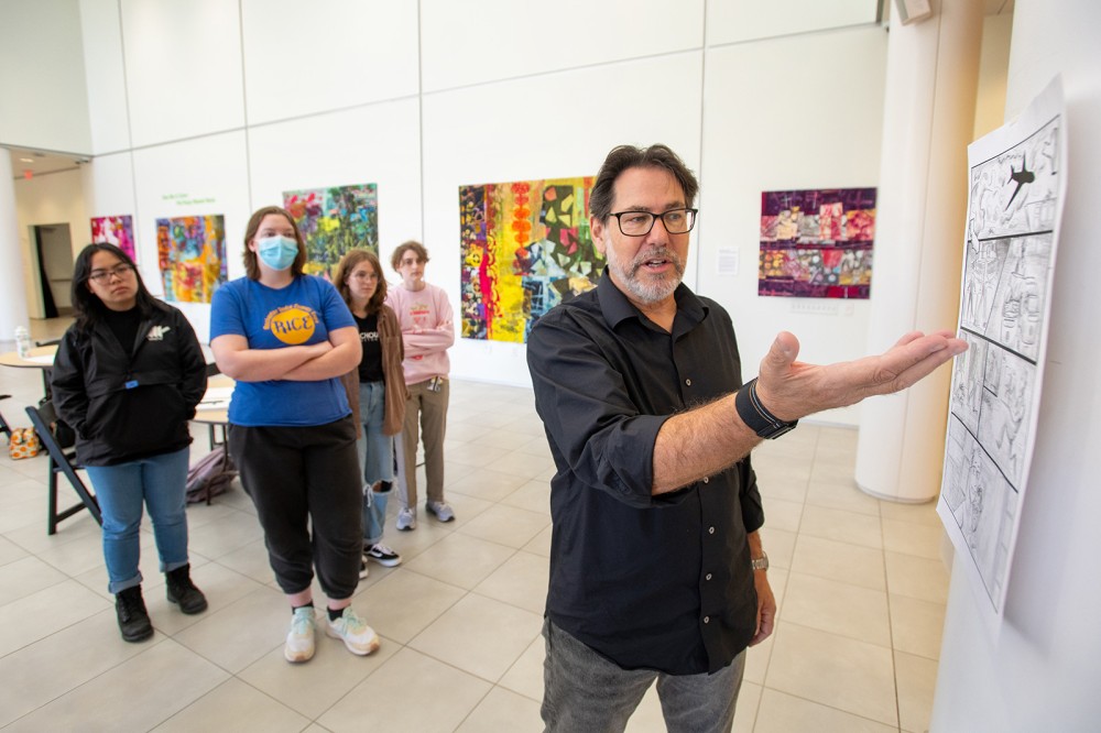 Adam Kubert stands in front of 4 students facing away from them. He has his arm raised palm-up, gesturing toward a page from a black and white comic that is taped to a wall in the University Gallery. Everyone in the group is looking at the comic page as Kubert speaks during a workshop.