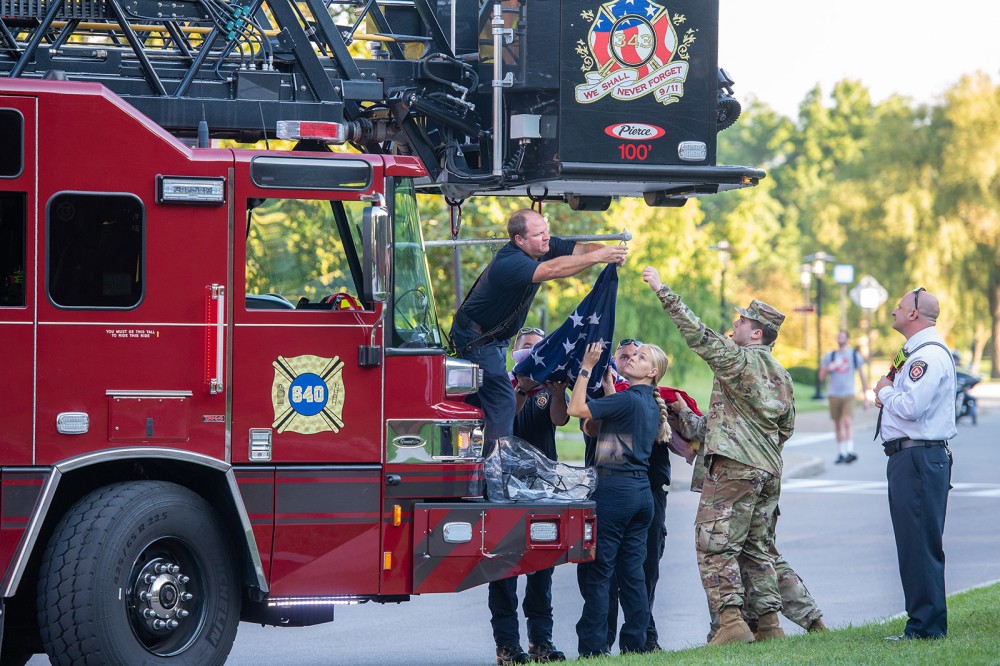 R O T C and fire department members attaching an American flag to the bucket of a ladder on a fire truck.