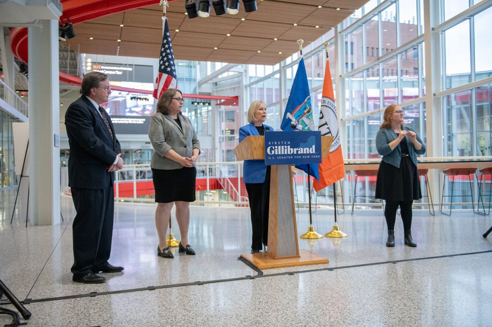 U S senator standing at a podium, with a sign language interpreter standing to her right and two college administrators standing to her left.