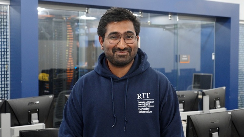 Student in nave blue hoodie with RIT logo and glasses stands in computing lab