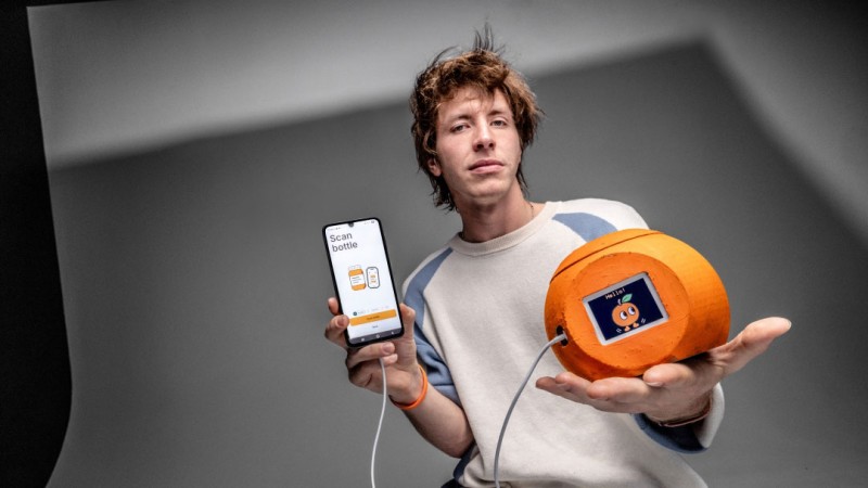 Student demonstrates a prototype consisting of a smartphone app and a connected orange, cylindrical device with a digital display. The smartphone screen shows a bottle-scanning interface, while the device displays a cartoon-style graphic. The student is seated in a studio setting with a neutral gray background, holding the phone in one hand and the prototype in the other.
