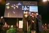 College student wearing graduation regalia on stage with two faculty members placing the ceremonial hood on her shoulders.