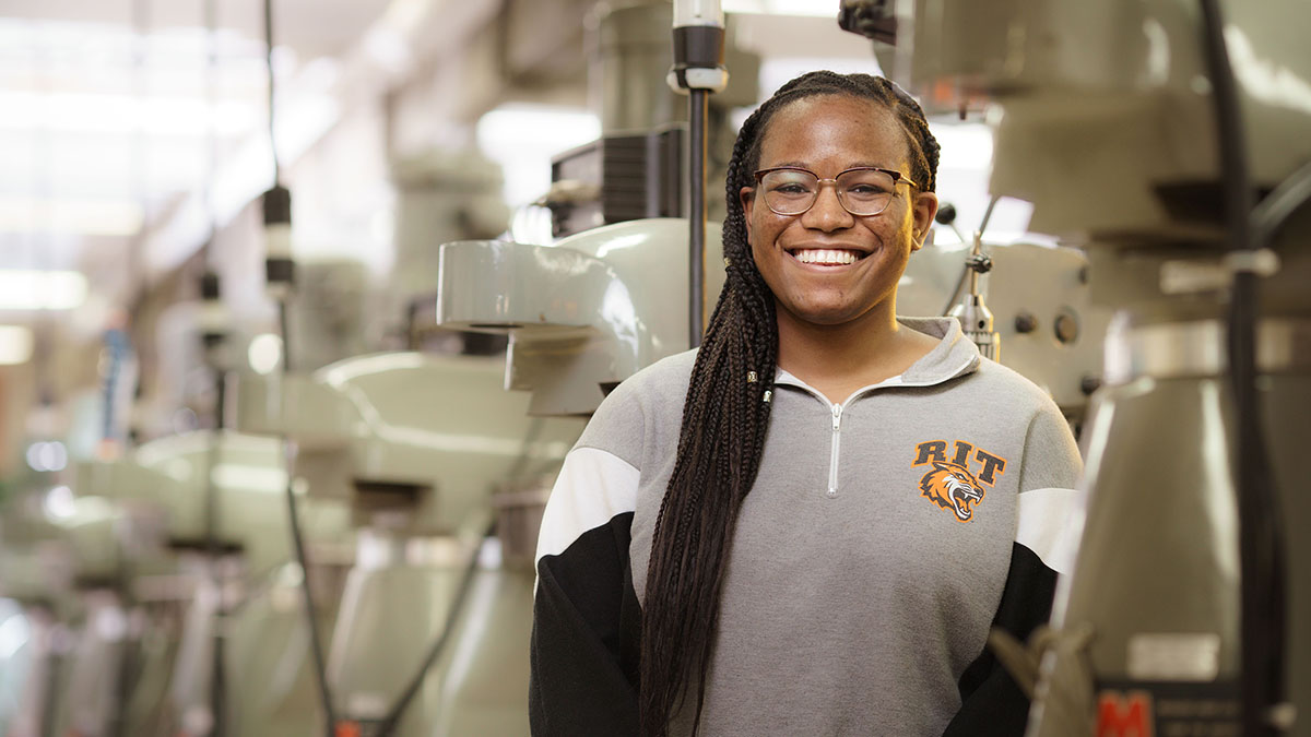 A smiling student wearing R I T apparel stands in a machine shop, surrounded by industrial equipment.