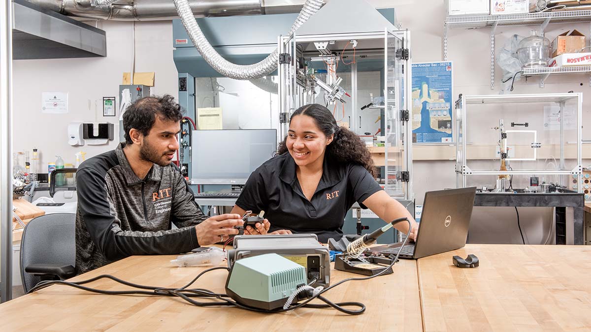 Two RIT students collaborate in a lab setting with electronics and a laptop on a workbench.