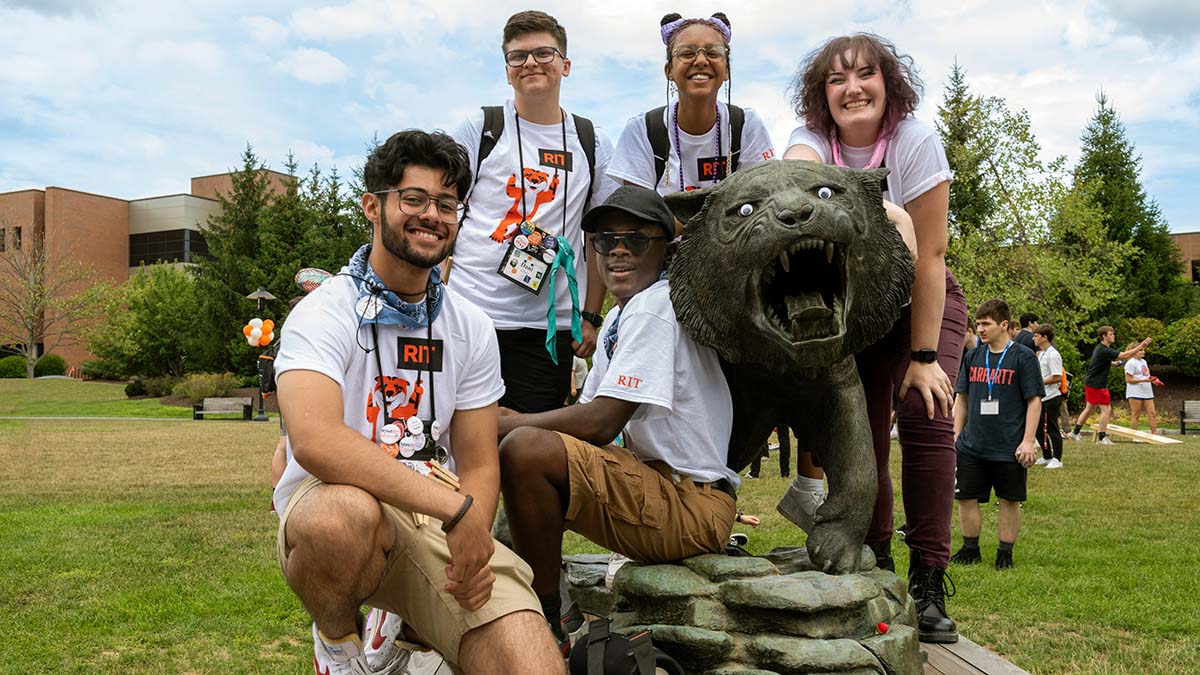 A group of smiling R I T students in university shirts pose together around a statue of a roaring tiger.