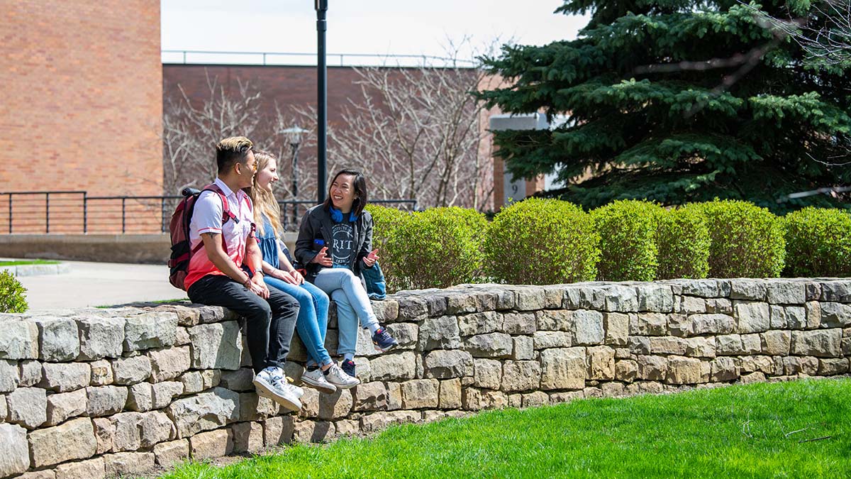 A group of students sits on a stone wall outdoors, engaging in conversation on a sunny day.