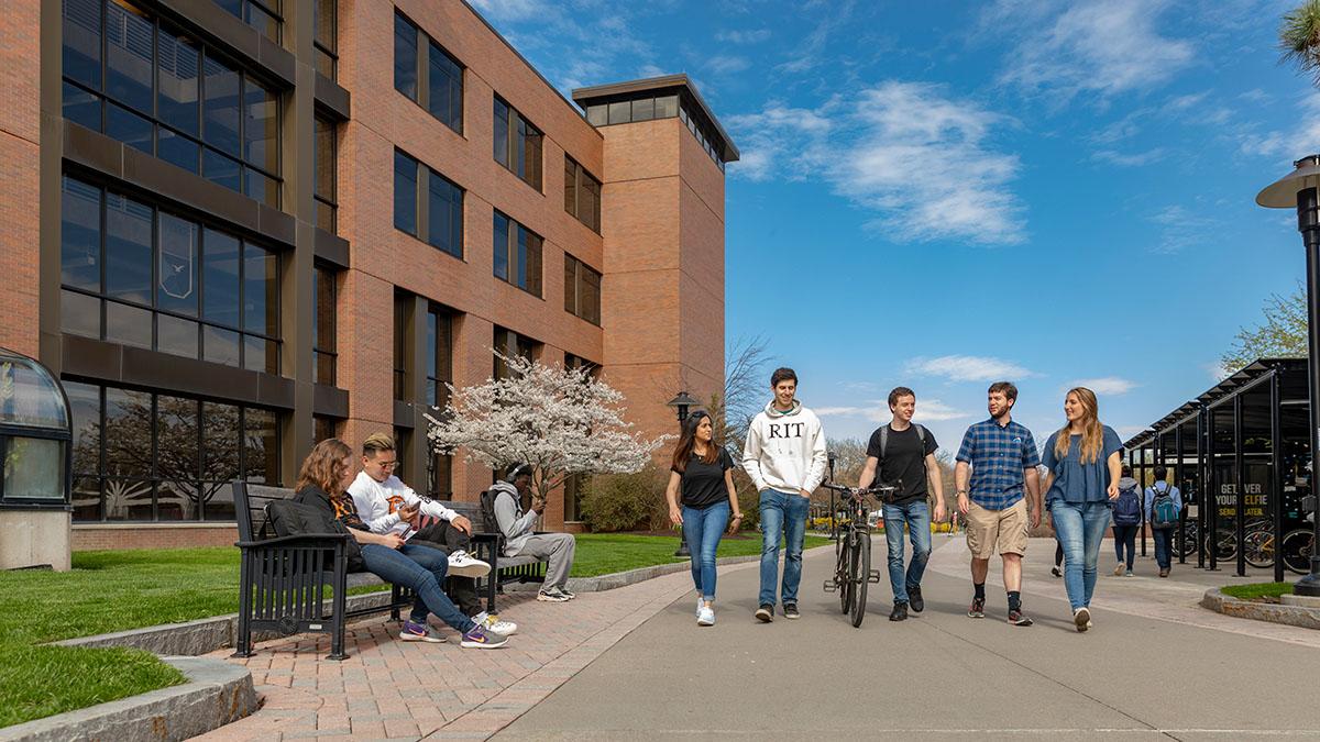 A group of students walking together on an R I T campus pathway, framed by modern brick buildings and springtime scenery.