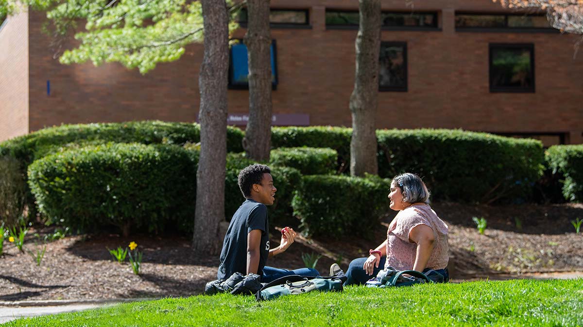 Two students sitting on a grassy area under trees, engaged in a friendly conversation near a campus building.