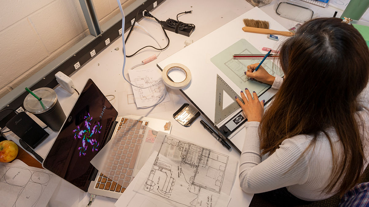 A student working on architectural sketches and designs at a cluttered desk, using rulers and pencils with open documents.