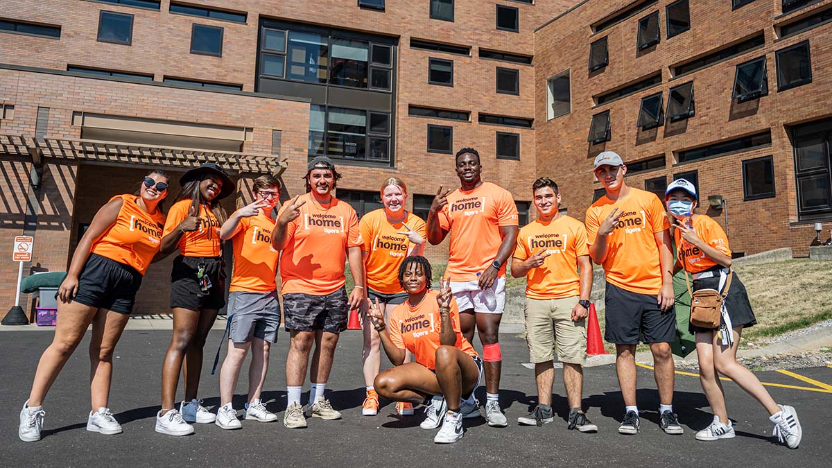 A group of students wearing matching Welcome Home Tigers shirts, posing happily in front of a residence hall.