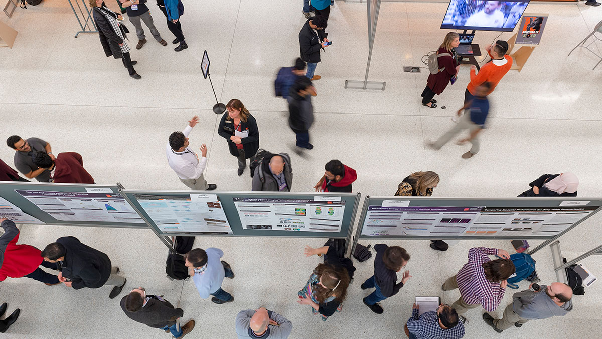 A large group of people mingling at a research poster session, with posters displayed on boards in a bright indoor venue.