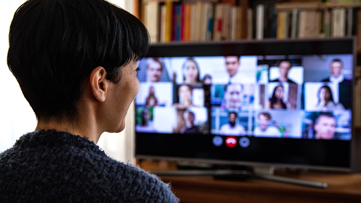 A person attending a virtual meeting with a video conferencing grid of participants displayed on a large screen.