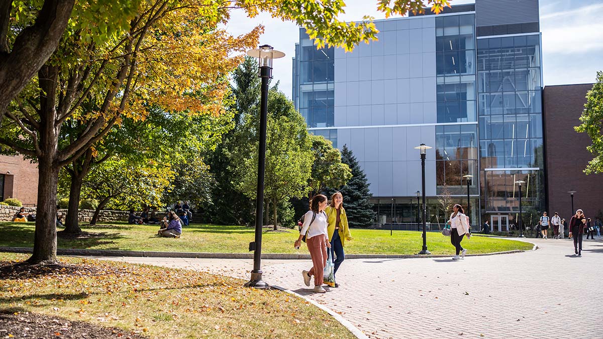 Students walking across a campus path in the fall, surrounded by trees with colorful leaves and modern academic buildings in the background.