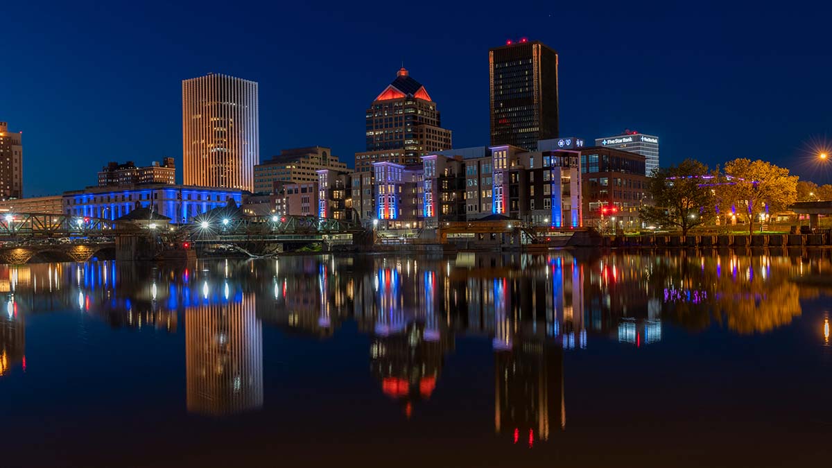 A vibrant night view of Rochester, New York’s skyline, with colorful lights reflecting on the calm river below.