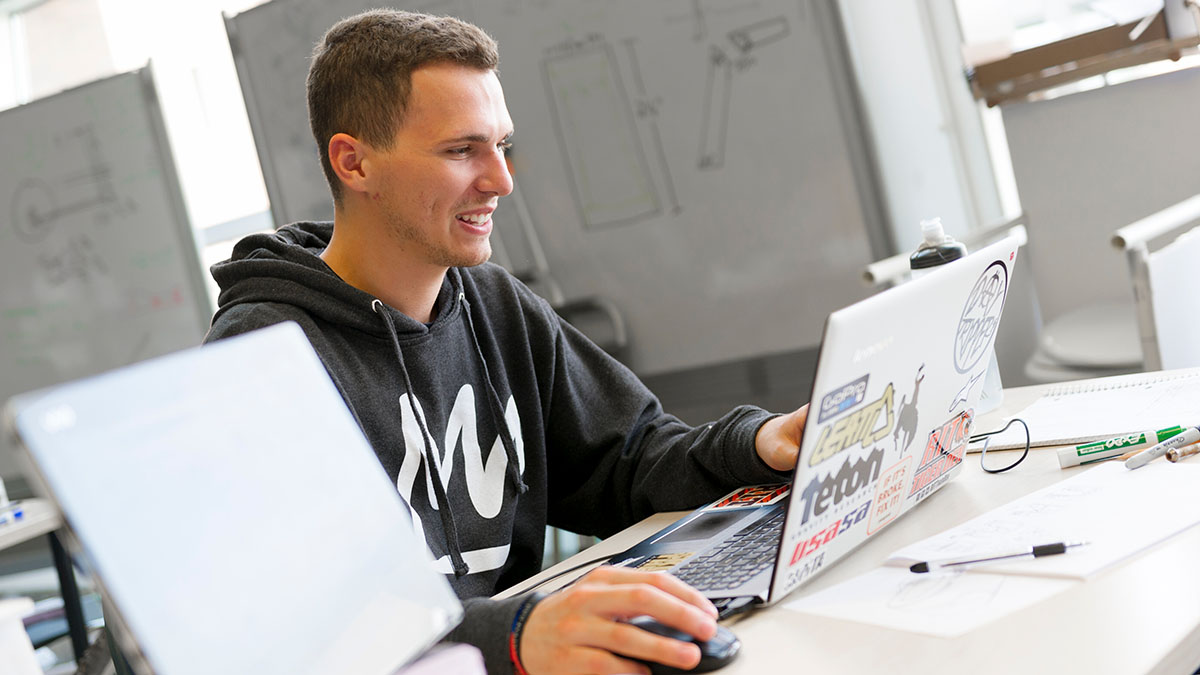 A student wearing a hoodie works on a laptop covered in stickers at a desk filled with papers.