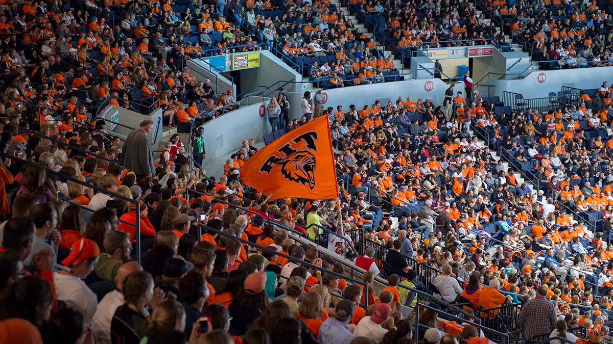 A large crowd of fans dressed in orange cheering during an R I T hockey game, with an R I T flag waving.