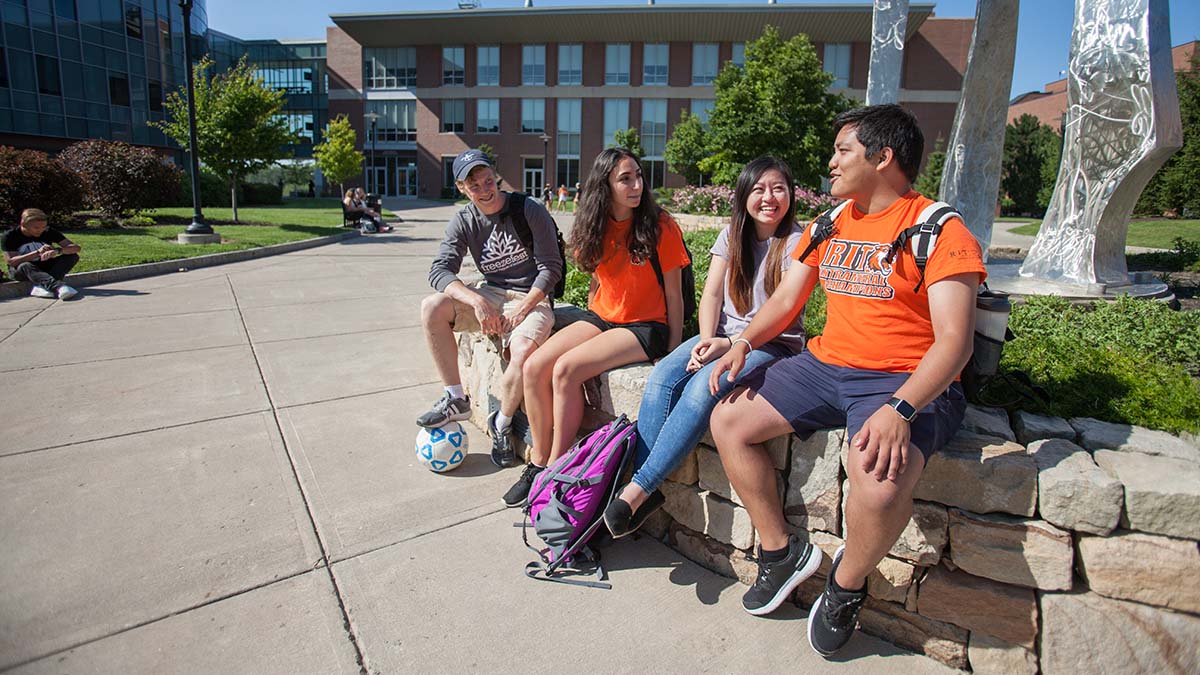 A group of students sitting outside on a sunny day, laughing and chatting near a campus sculpture.