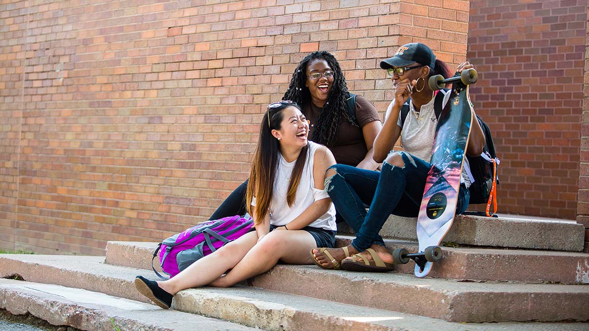 Three students sitting on steps outside, chatting and laughing together.