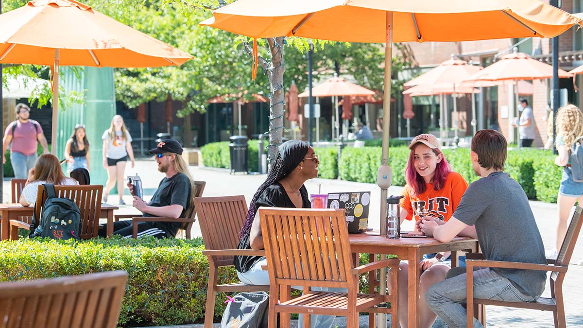 A group of students seated outside under umbrellas, engaging in conversation.