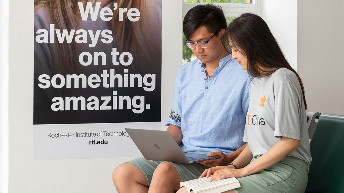 Two students sitting in front of a poster reading We're always on to something amazing, using a laptop together.