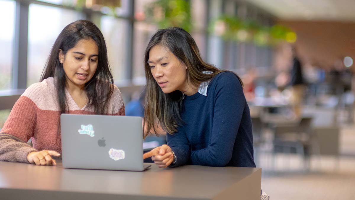 Two students collaborating on a laptop in a bright common area.