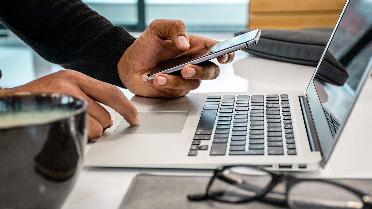 A person holding a phone in one hand and working on a laptop with the other.