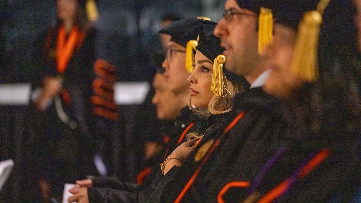 Graduates in black caps and gowns with gold tassels sitting in a ceremony.