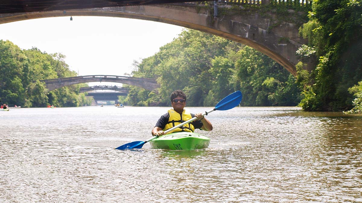 A person kayaking on a river with bridges and greenery in the background.