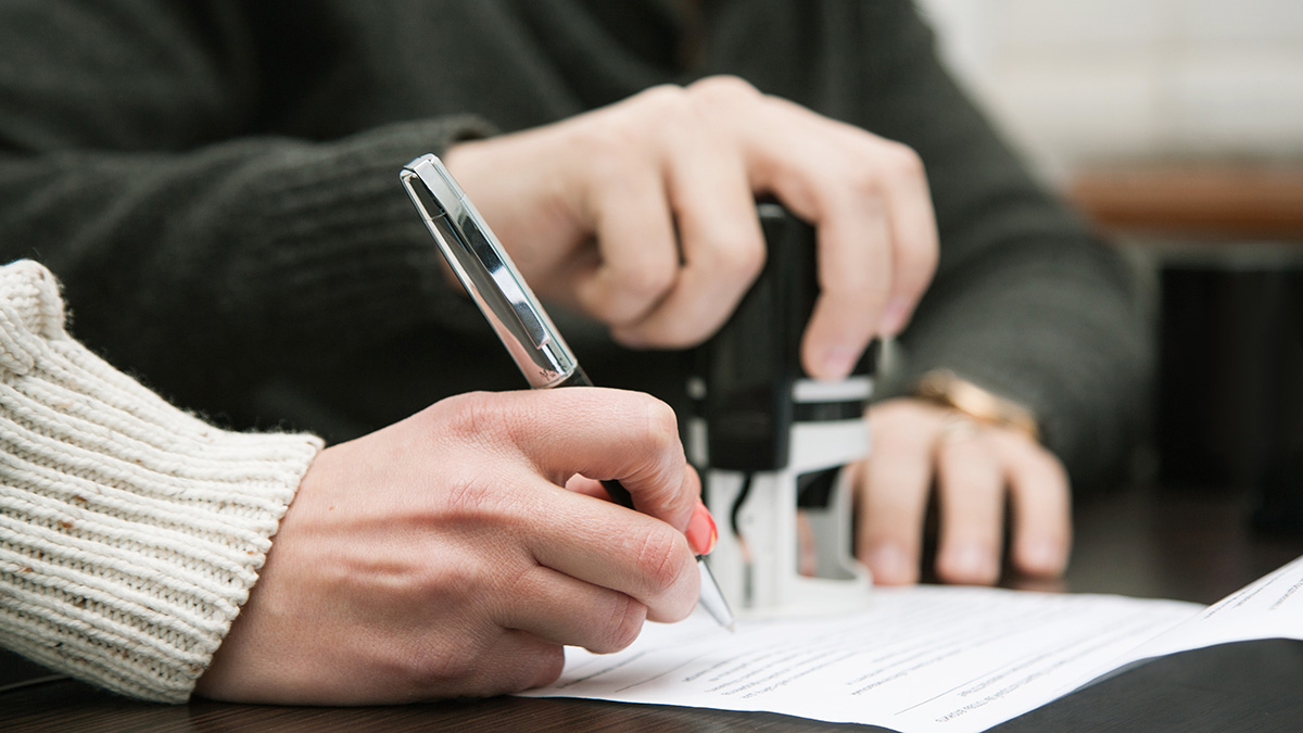 A close-up of hands stamping and signing a document on a desk.