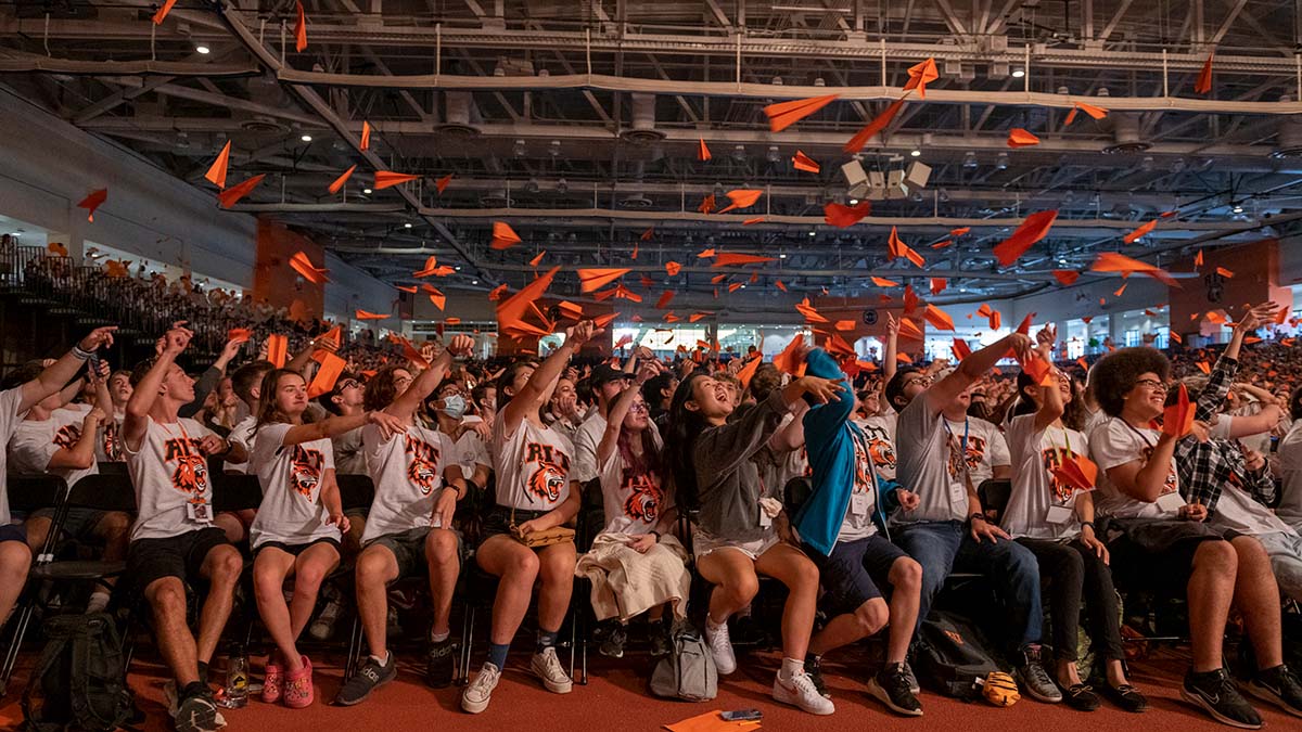 Students enthusiastically throwing orange paper airplanes in the air.