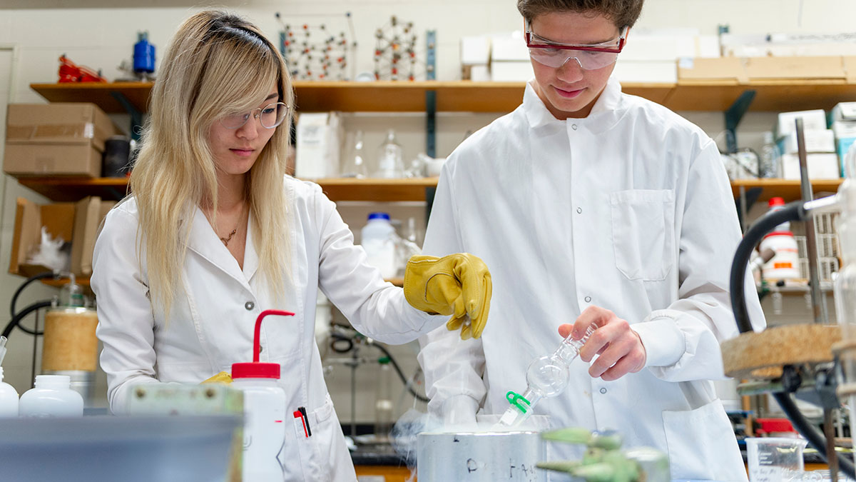 Two students in lab coats and goggles conducting an experiment in a chemistry lab.