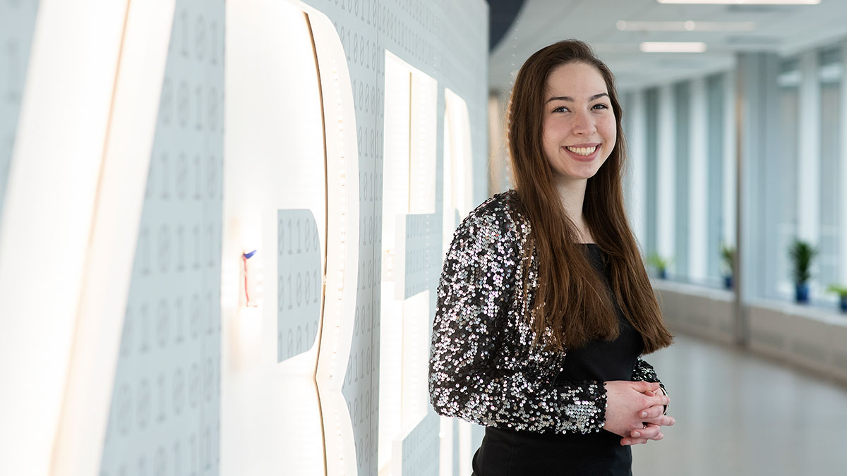 A student smiling in front of a brightly lit installation with binary code in the background.