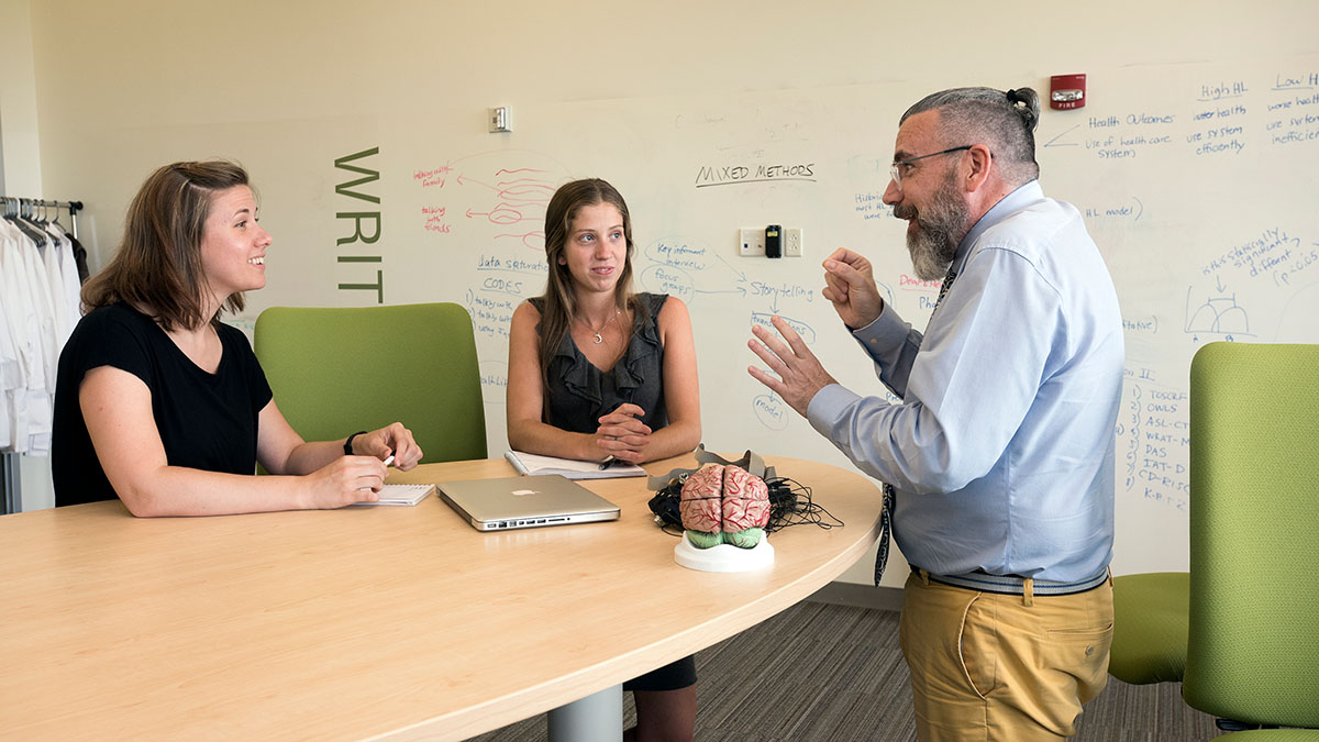 Three people in discussion, one using sign language, at a round table with notes and a model brain on display.