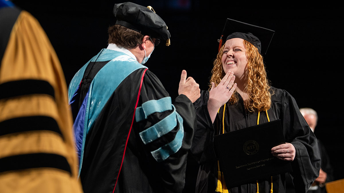 A graduate in a cap and gown smiling and signing thank you while receiving a diploma on stage.