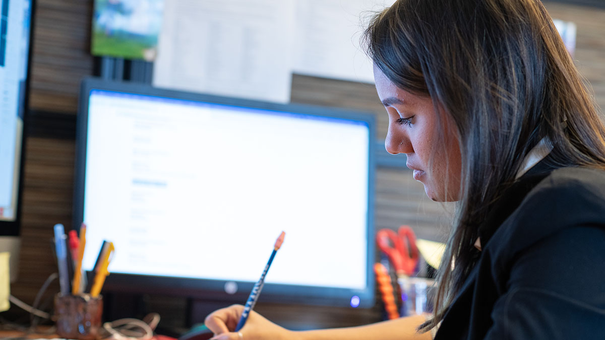 A focused employee writing with a pencil while working at a computer desk with a monitor in the background.