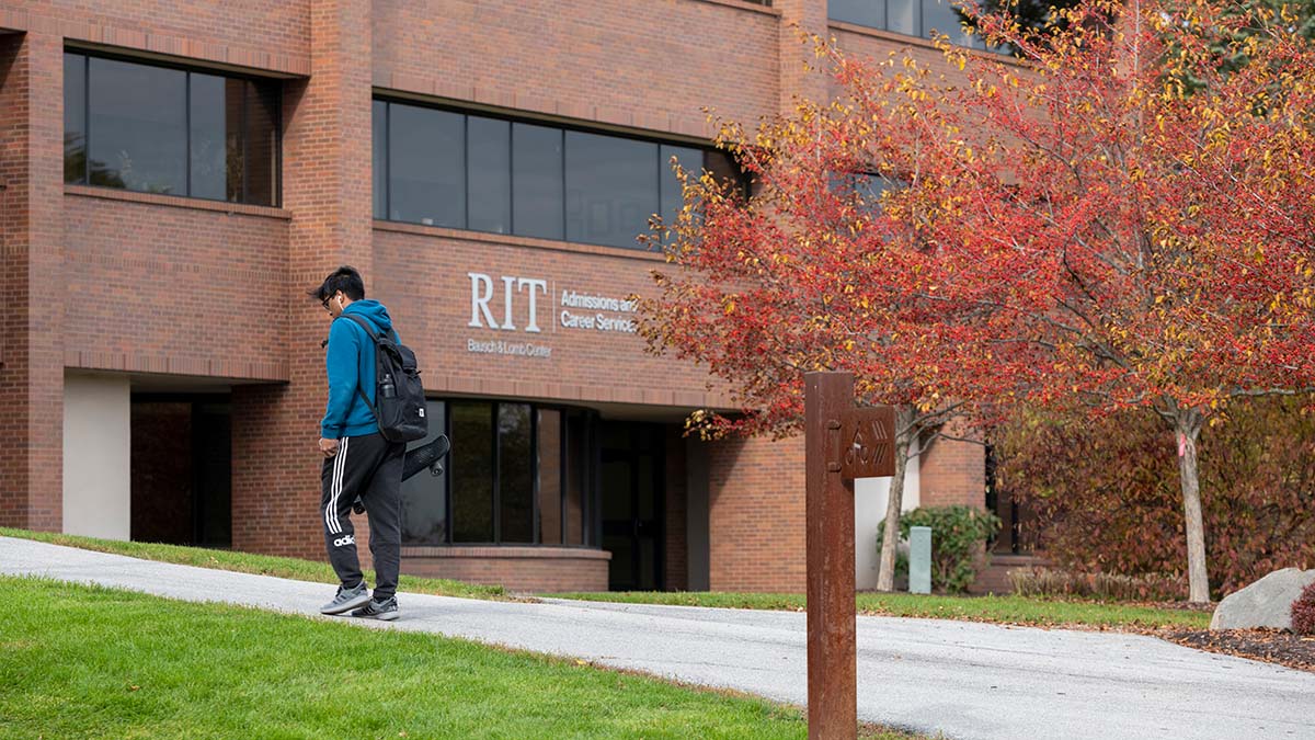 A student walking by the R I T Bausch and Lomb Center with autumn foliage in the background.