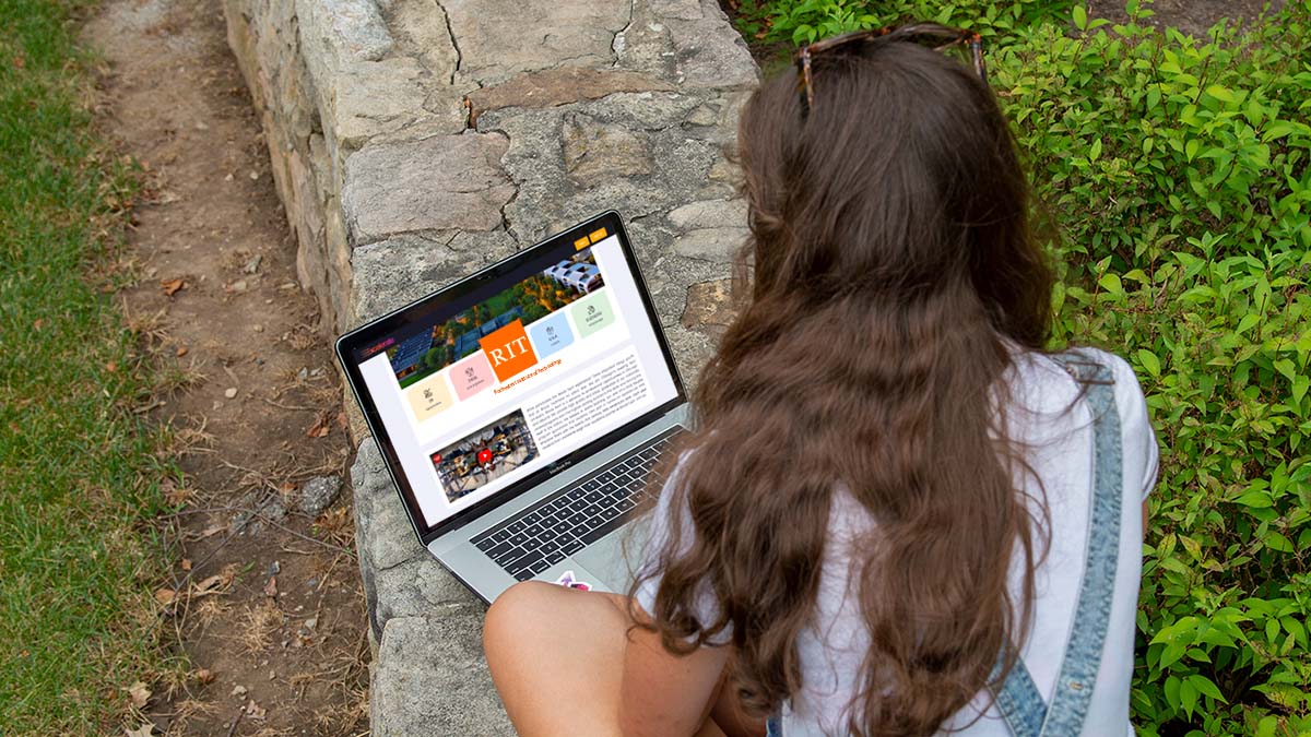 A student browsing the R I T website on a laptop outdoors on a stone wall.