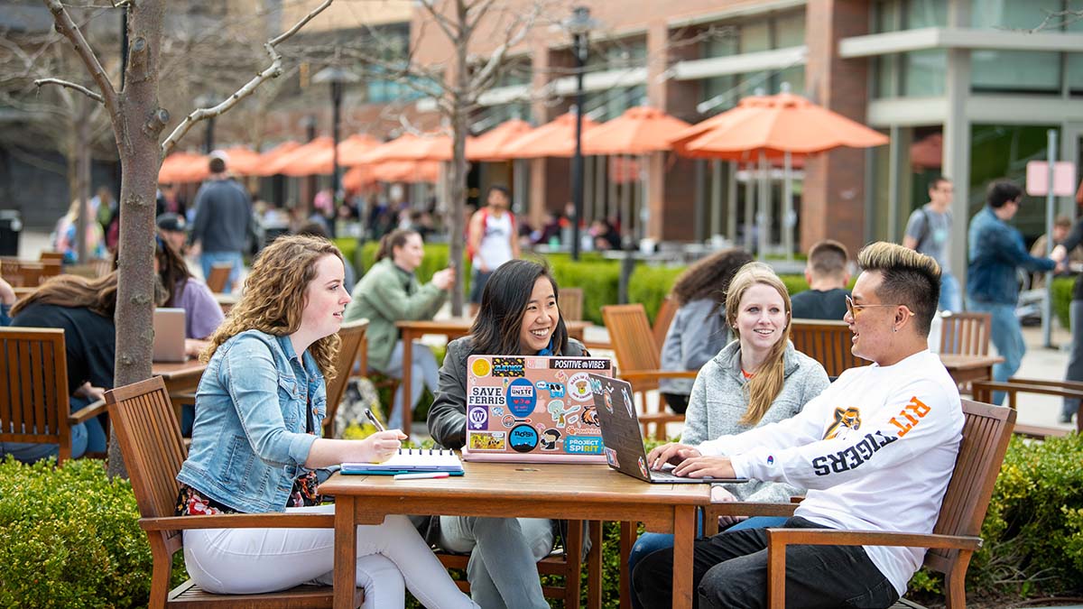 A group of students enjoying time outdoors at wooden tables with laptops and notebooks on a sunny day.