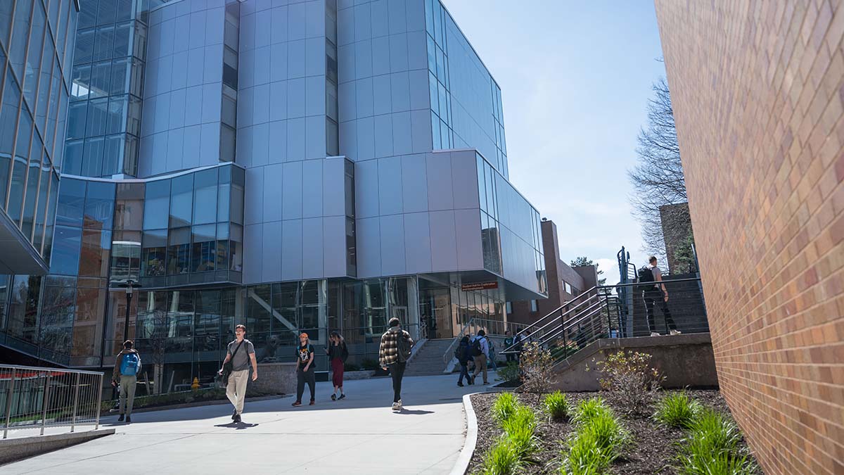 Students walking near a modern glass and metal building on the R I T campus under a clear sky.